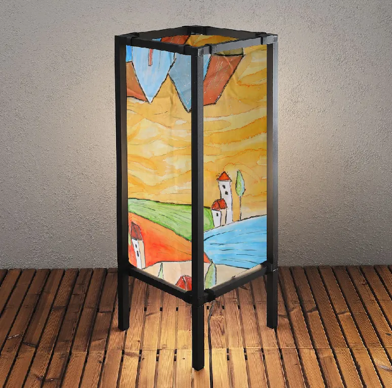 A tall square lamp with a black frame and painted panels showing an ochre sky, green and orange fields, a blue river and small red-roofed houses.