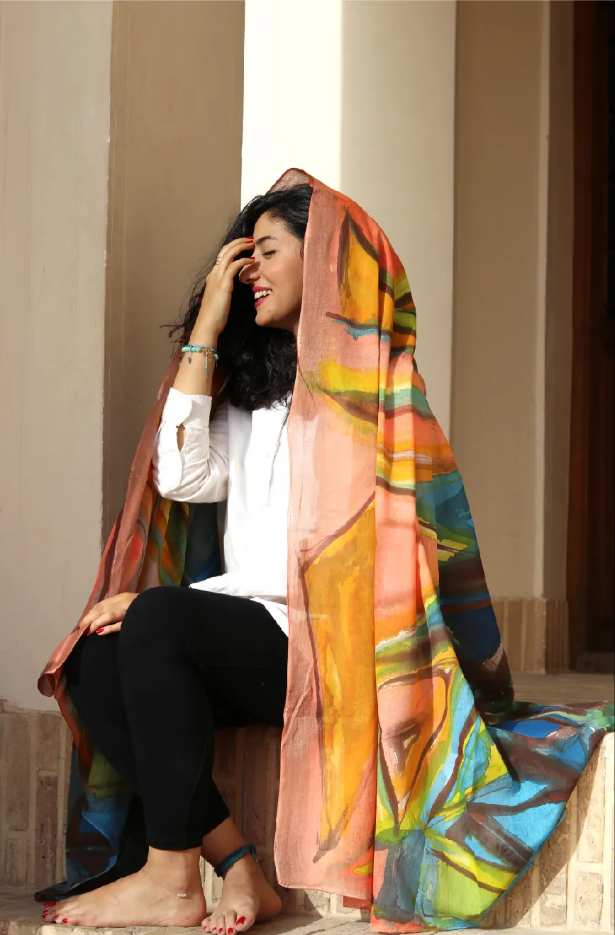 A woman sitting on a step, laughing, with a hand-painted wrap in coral, orange, gold and blue draped over her head and falling to the floor.