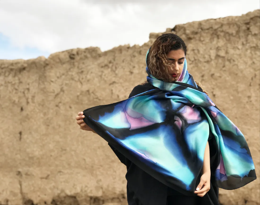 A woman in front of an old mud-brick wall lifts a hand-painted scarf in black, deep blue, turquoise and soft pink.