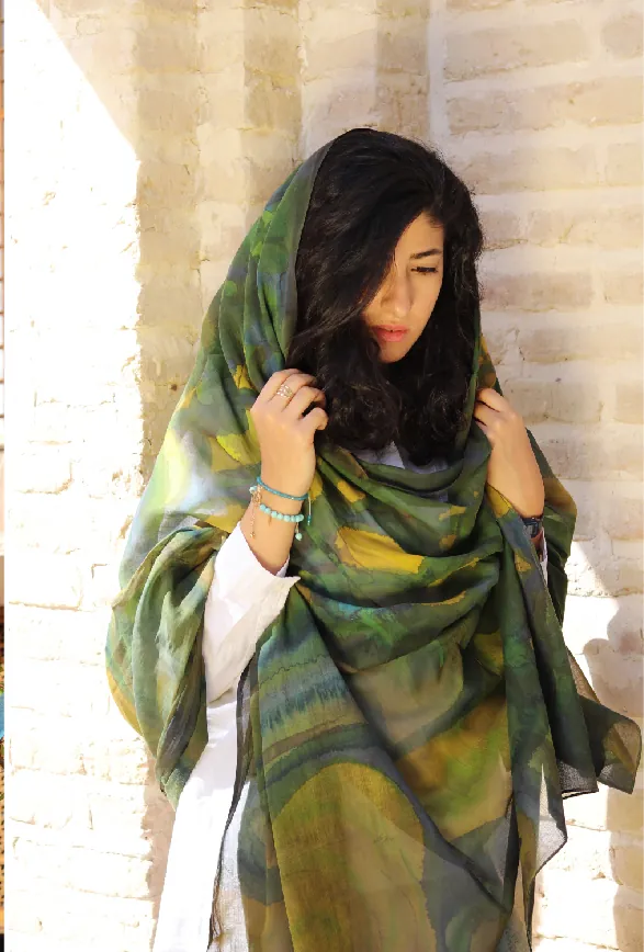 A woman leaning against a pale brick wall wears a hand-painted wrap in olive green, ochre and gray over her hair.