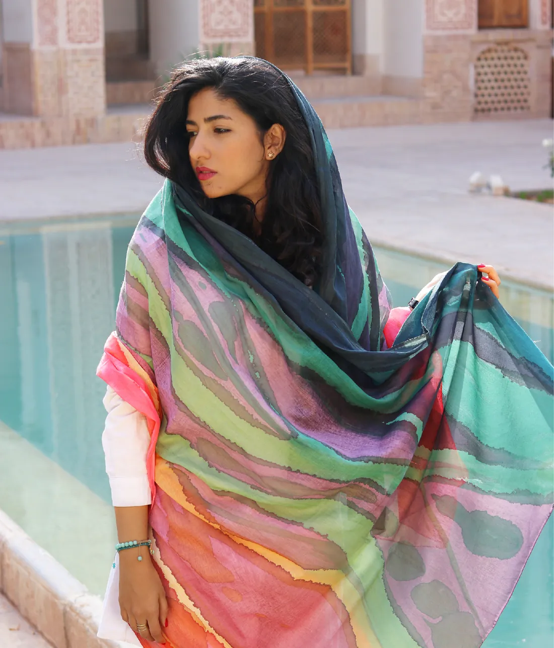 A woman beside a courtyard pool wears a large hand-painted wrap over her head and shoulders, painted in wavy bands of teal, violet, lime, coral and gold.