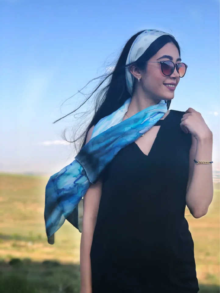 A woman in sunglasses outdoors, with a hand-painted scarf in sky blue and white tied over her hair and around her shoulders.