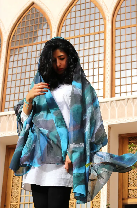 A woman in front of tall arched lattice windows wears a hand-painted wrap in turquoise, slate blue and gray around her head and shoulders.