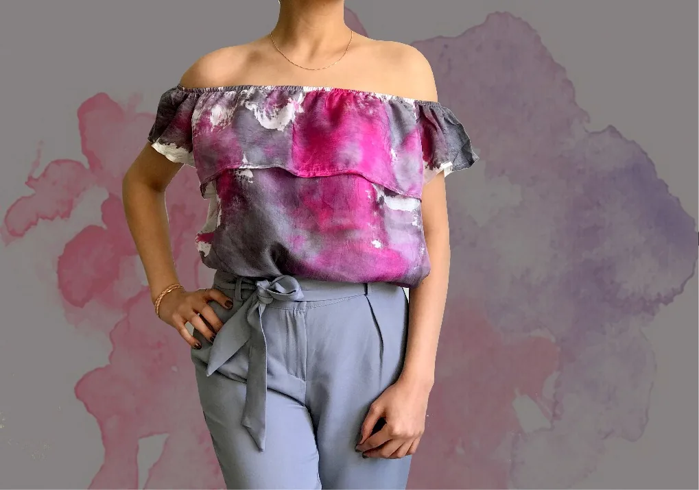 An off-shoulder ruffled top painted in magenta, violet and charcoal gray, worn with gray-blue trousers.