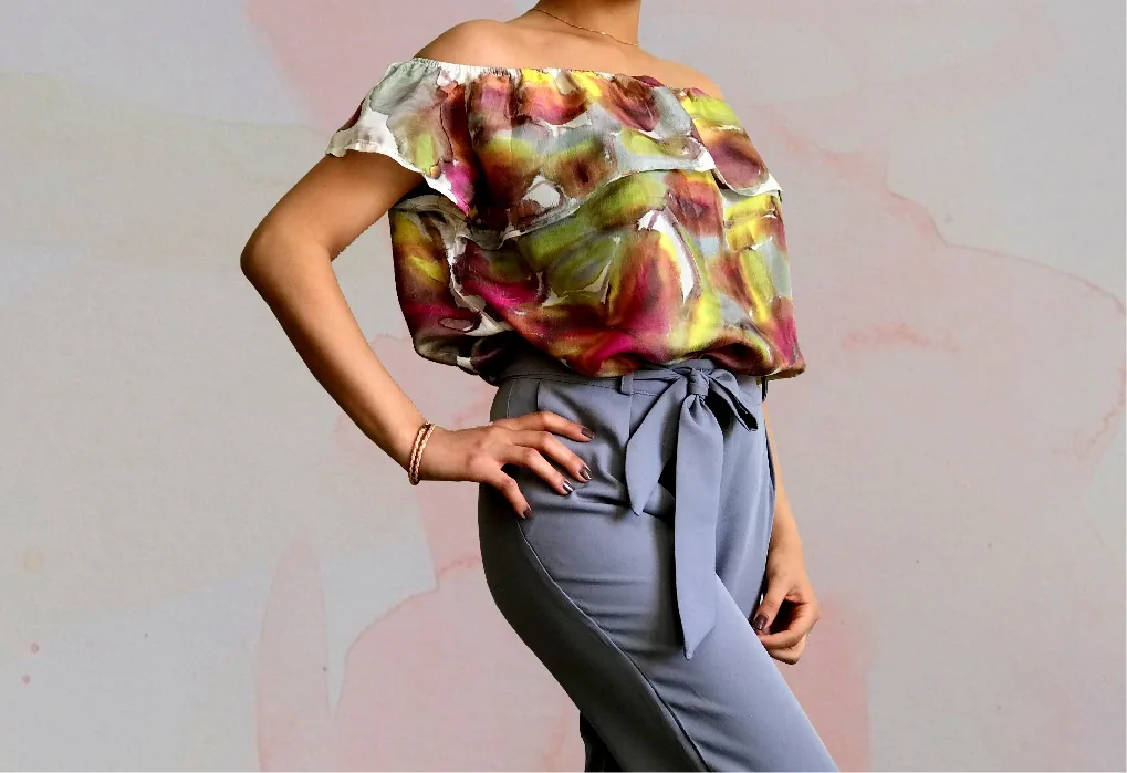 An off-shoulder ruffled top painted in rose, lime and gray, worn with gray-blue trousers tied at the waist.