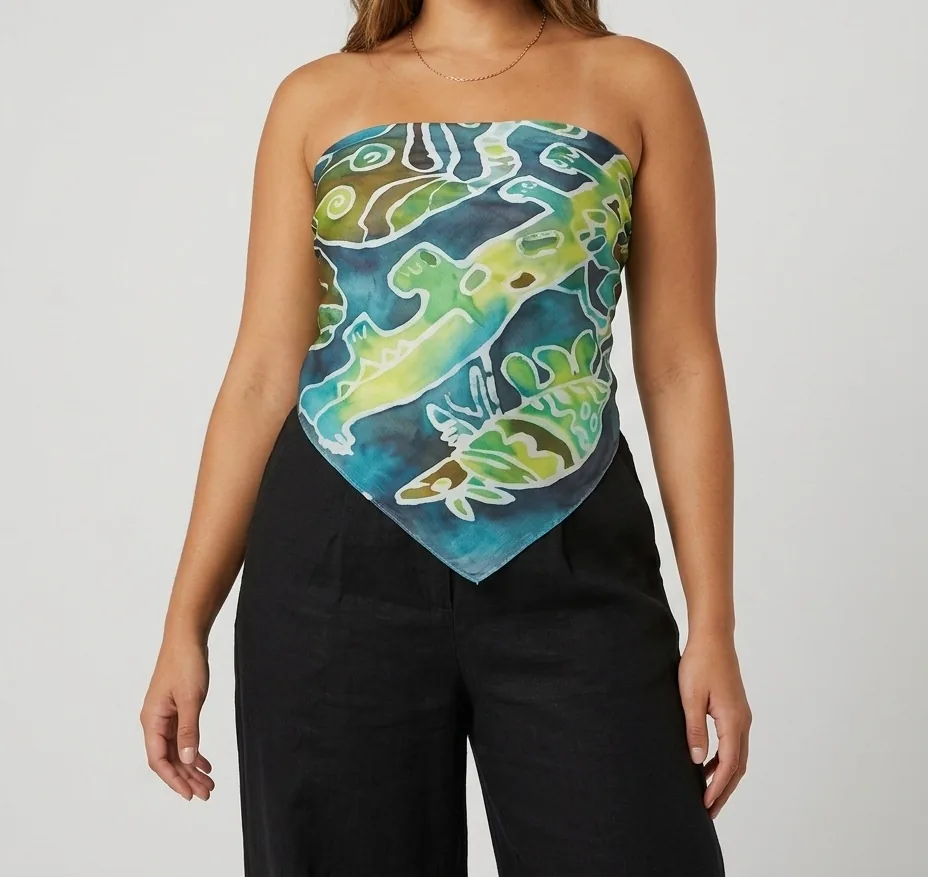 A woman wearing a hand-painted silk scarf tied as a strapless top. Lime and yellow-green lizards and leafy creatures, outlined in pale resist lines, swim across deep teal and blue.